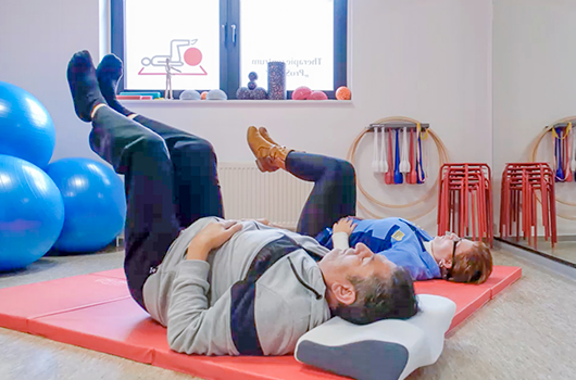 Auf dem Bild siehst du eine Therapeutin mit ihrem Patienten Übungen in Rückenlage auf der Bodenmatte praktizieren.