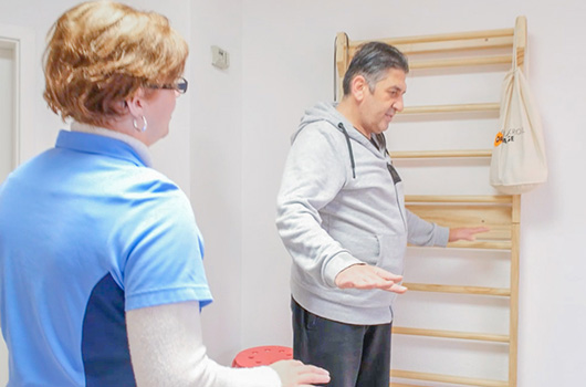 Das Bild zeigt einen Patienten bei einer Balanceübung im aufrechten Stand, die von der Ergotherapeutin angeleitet wird.